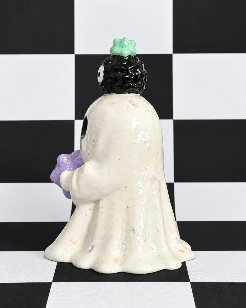 A ceramic ghost figurine with a speckled cream body, black hair, a green flower, and a purple flower, standing on a black and white checkered background.