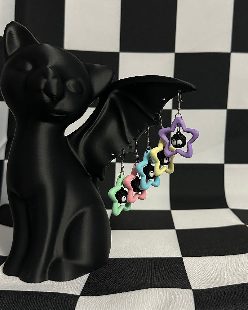 Black bat-shaped figurine with colorful star-shaped earrings on a black and white checkered background