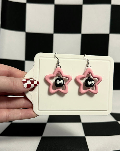 Pink star-shaped earrings on a card held by a hand against a black and white checkered background