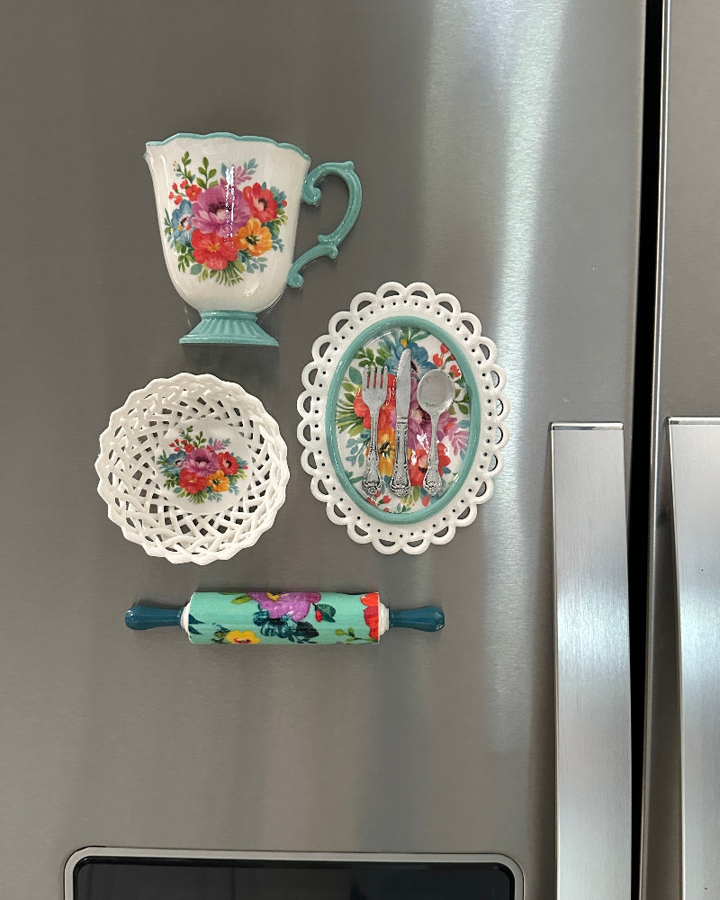 Decorative magnets shaped like a teacup, plate, and rolling pin on a refrigerator.