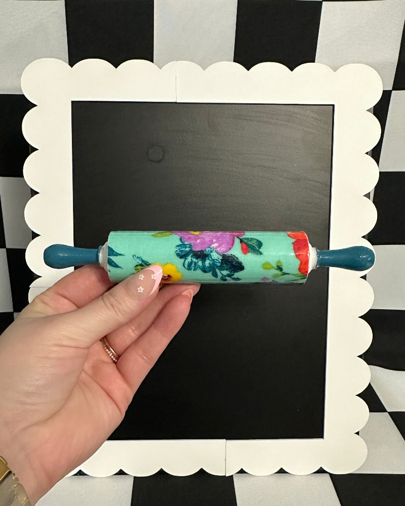 Hand holding a floral-patterned rolling pin against a black and white checkered background
