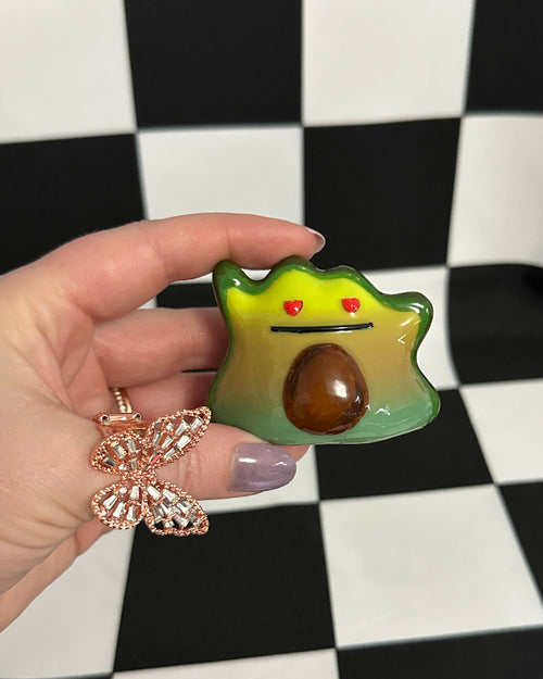 Hand holding a small green avocado-shaped object with a brown center against a black and white checkered background.