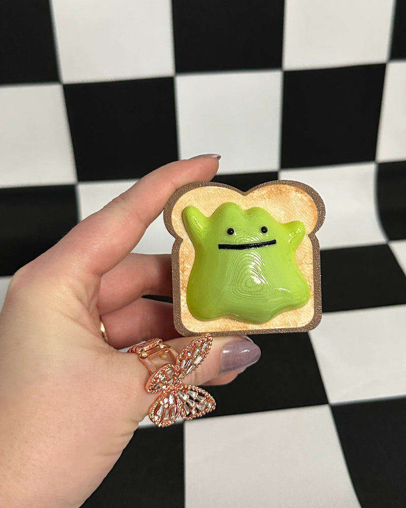 Hand holding a small green creature on a piece of bread against a black and white checkered background