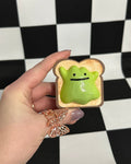 Hand holding a small green creature on a piece of bread against a black and white checkered background