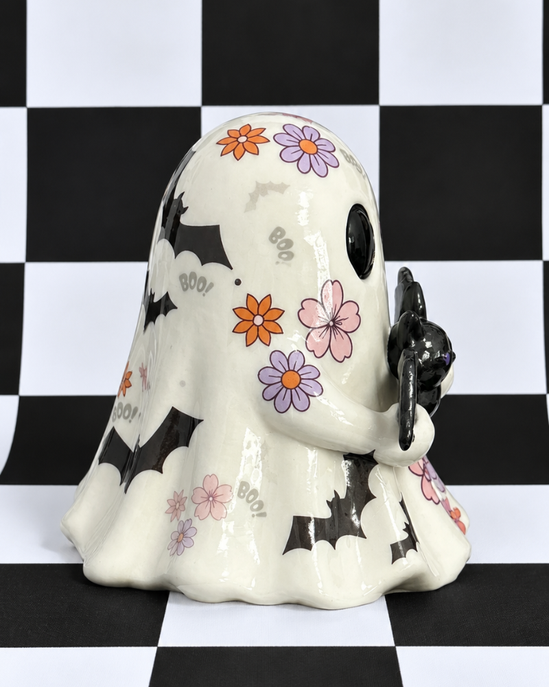A ceramic ghost figurine decorated with black bat silhouettes, orange and purple flowers, and pink blossoms, holding a black cat. The figurine is set against a black and white checkered background.