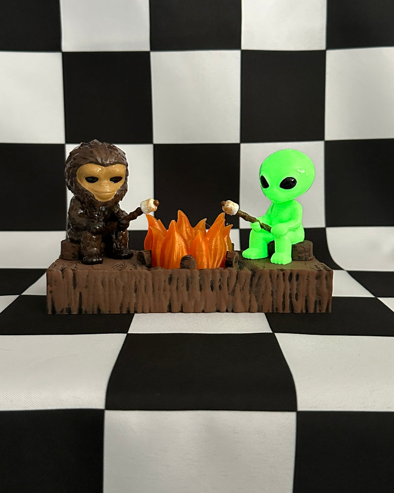 Two figurines, one resembling a creature and the other a green alien, are sitting around a campfire on a black and white checkered background.