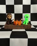 Two figurines, one resembling a creature and the other a green alien, are sitting around a campfire on a black and white checkered background.