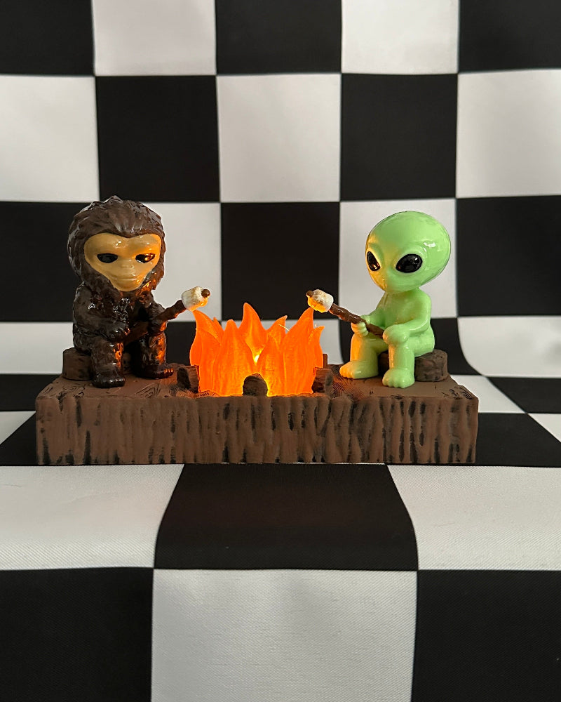 Two figurines, one resembling a human and the other an alien, are sitting around a campfire on a checkered black and white background.