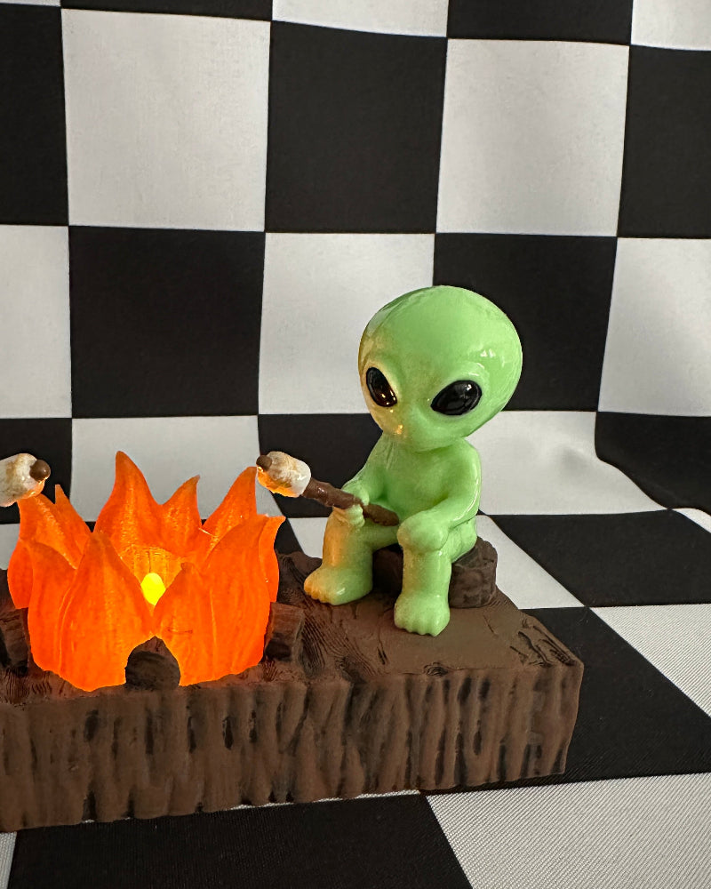 Green alien figurine roasting a marshmallow over a campfire on a checkered background