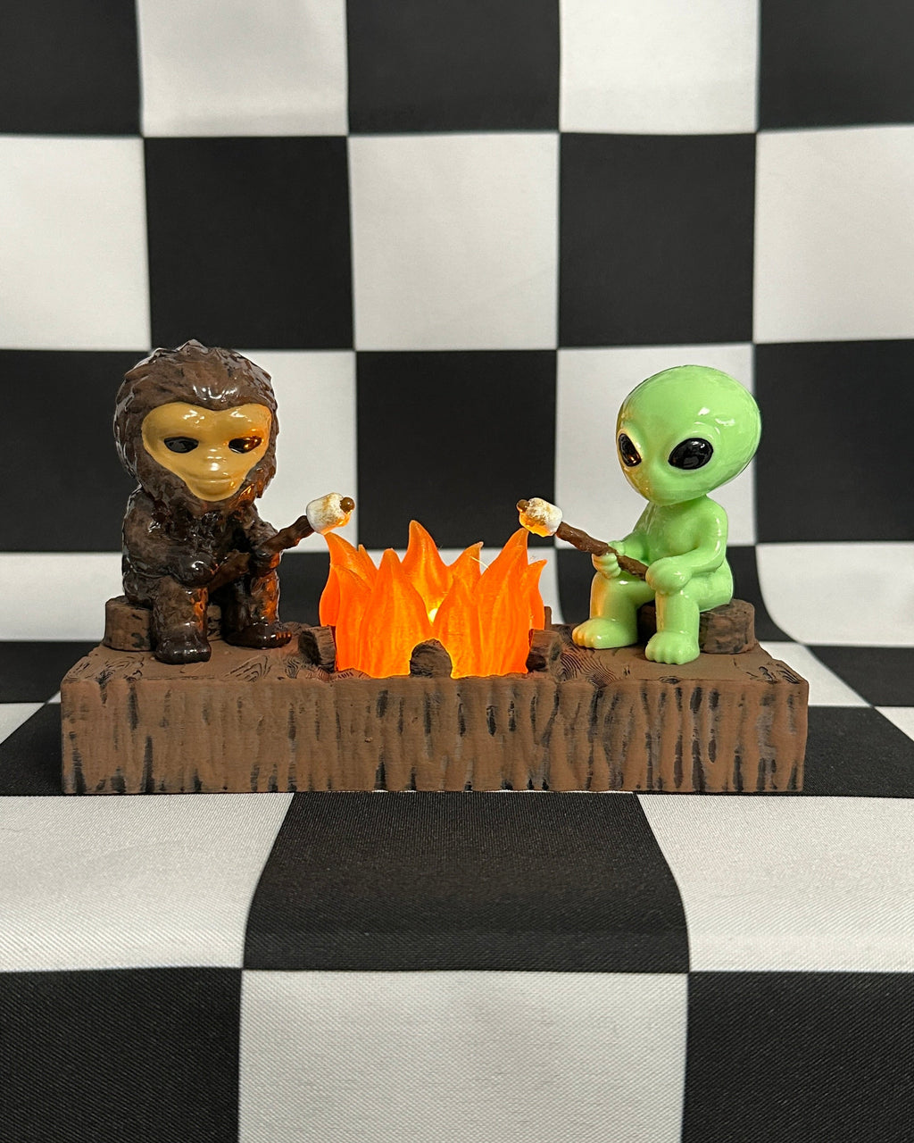 Two figurines, one resembling a human and the other an alien, sitting around a campfire on a checkered background.
