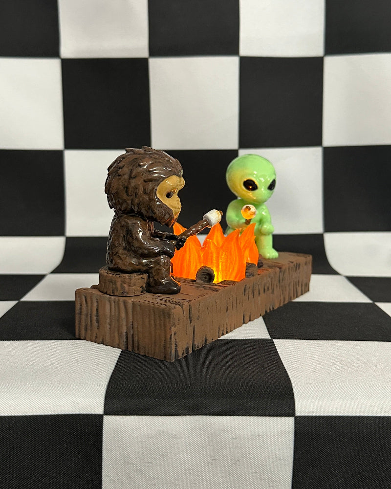 Two small figurines, one resembling a creature and the other an alien, sitting around a campfire on a checkered background.