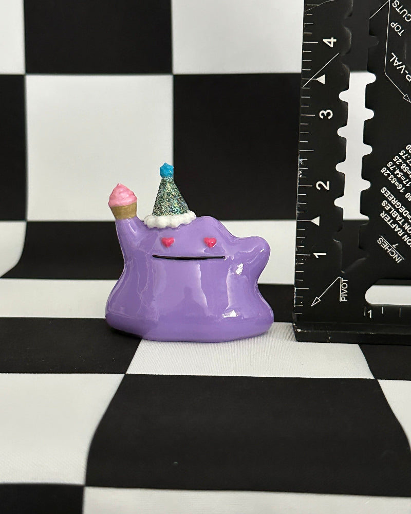 Purple creature figurine with a blue hat and pink cheeks on a checkered background with a ruler for scale.