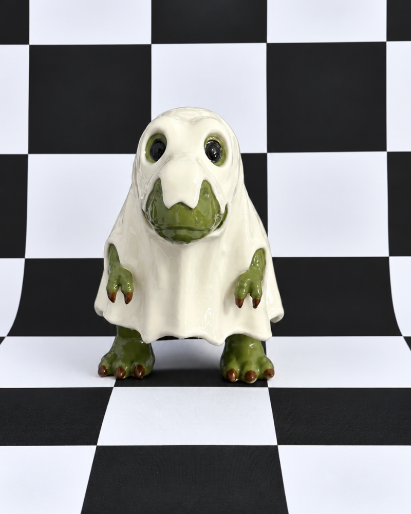 A small ghost-shaped figurine or toy with a white body and green face, hands, and feet, standing on a black-and-white checkered studio background.