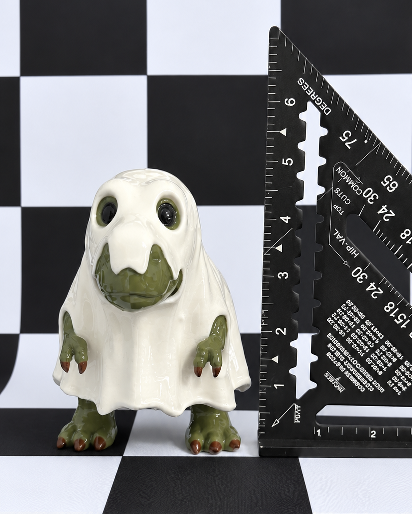 A small ghost-shaped figurine with green skin and a white sheet-like body stands on a black-and-white checkered surface next to a black triangular ruler.
