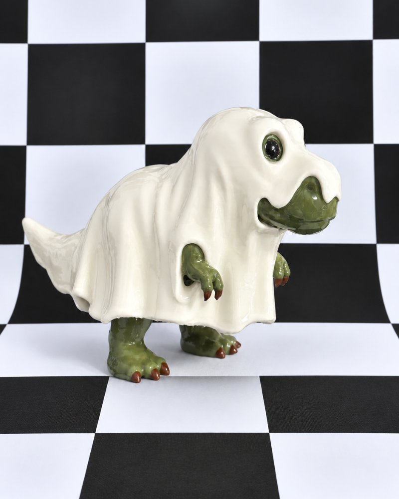 A small green dinosaur figurine draped in a white ghost-like sheet, standing on a black-and-white checkered surface and backdrop.
