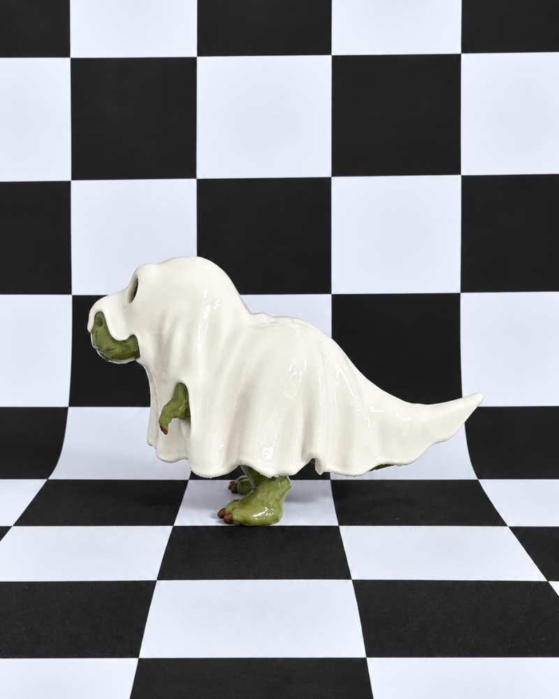 A small figurine or sculpture of a ghostly dog-like creature draped in a white sheet, with green feet and hands, standing on a black-and-white checkered floor and matching checkered backdrop.