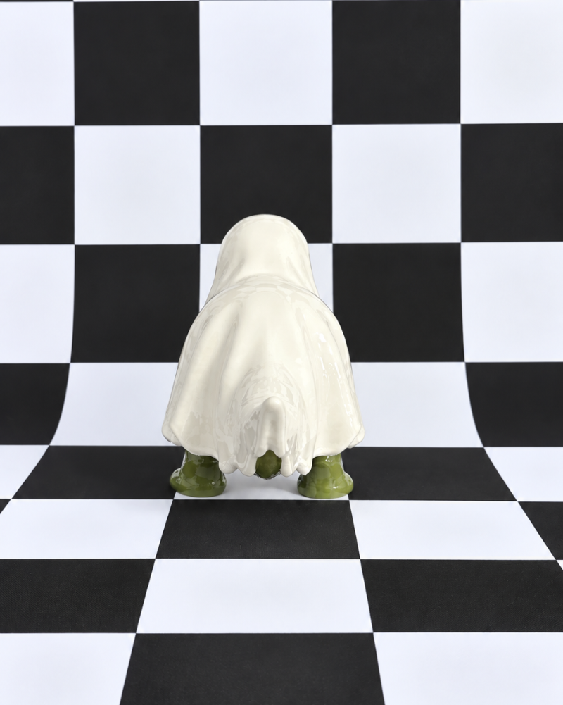 A small figurine or sculpture draped in a white sheet, viewed from behind, standing on a black-and-white checkered floor and matching backdrop.