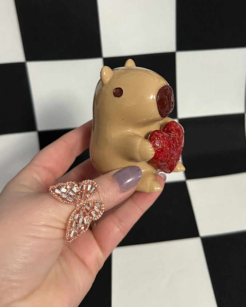 Hand holding a small figurine of a capybara with a heart against a checkered background