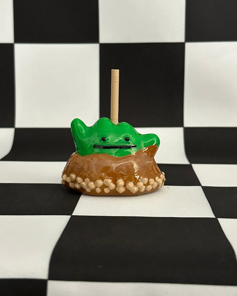 Green frog-shaped candy on a stick with a brown base against a black and white checkered background