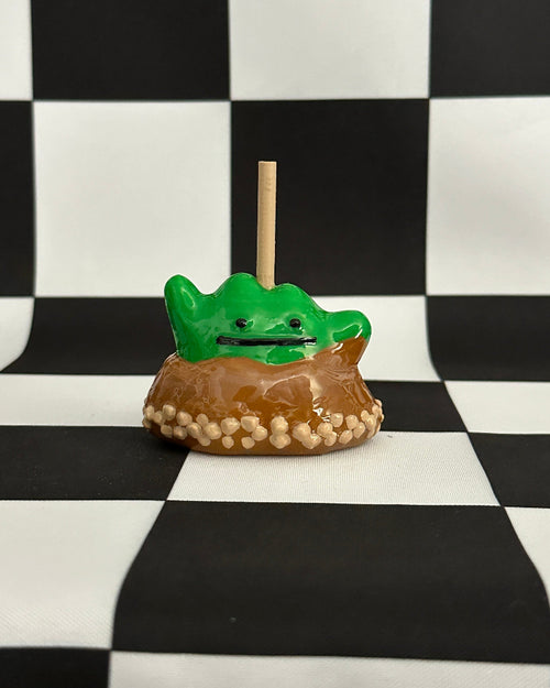 Green frog-shaped candy on a stick with a brown base against a black and white checkered background
