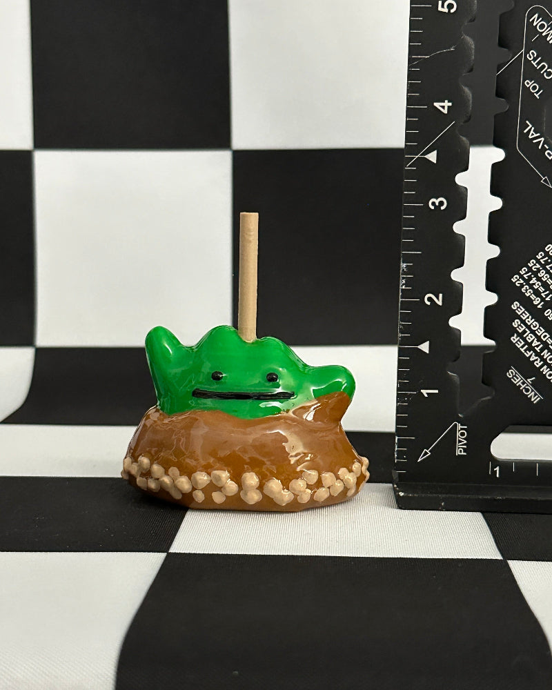 Small green frog figurine on a brown base with a ruler for scale on a checkered background