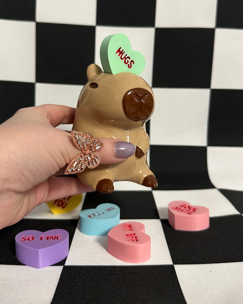 Hand holding a small capybara figurine with a green heart tag on a checkered background