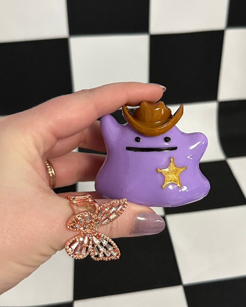 Hand holding a purple character figurine with a gold crown and star against a black and white checkered background