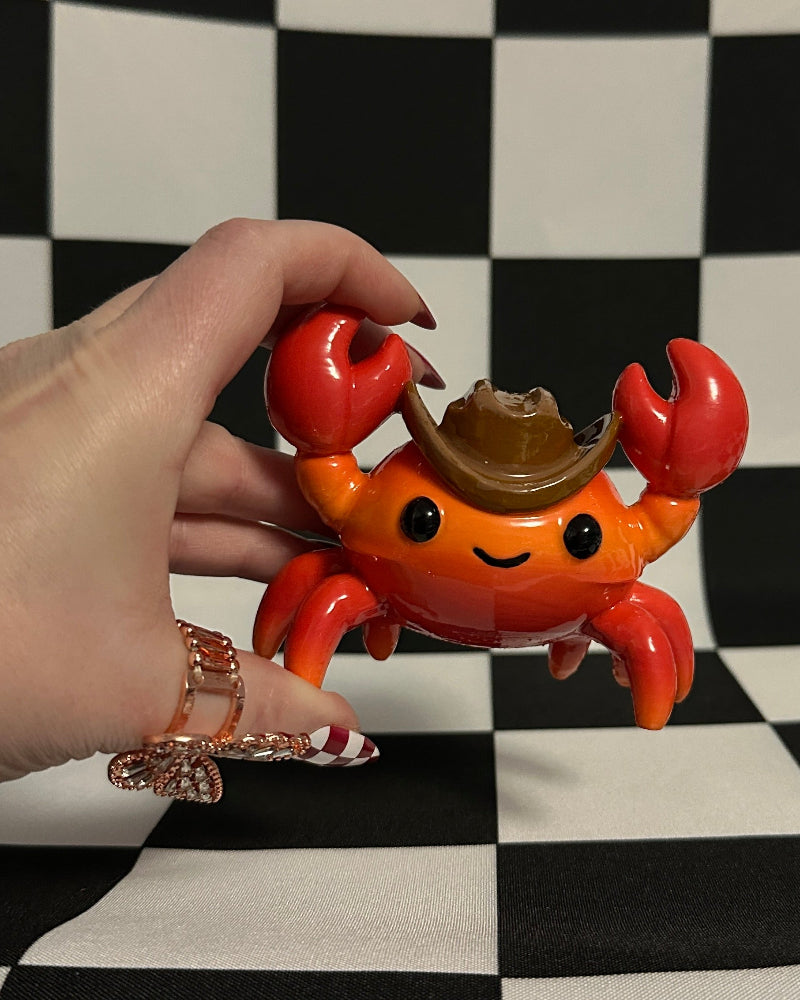 Toy crab figurine with a cowboy hat held by a hand on a black and white checkered background