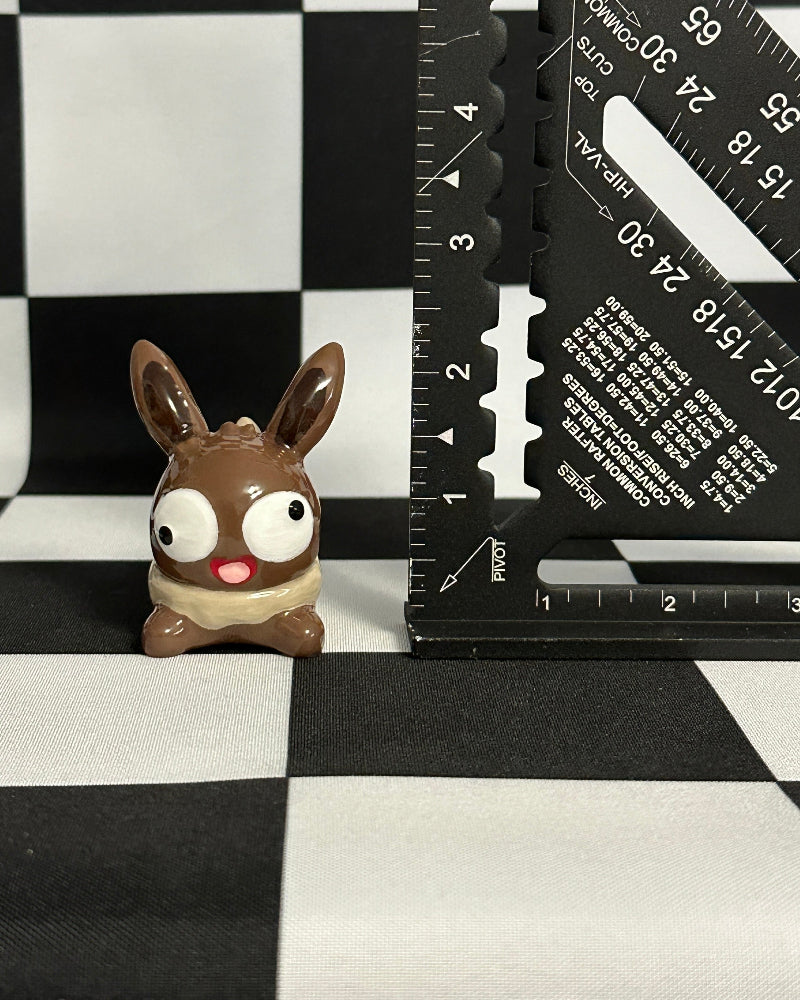 Small figurine of a brown creature with large eyes on a black and white checkered background with a ruler.