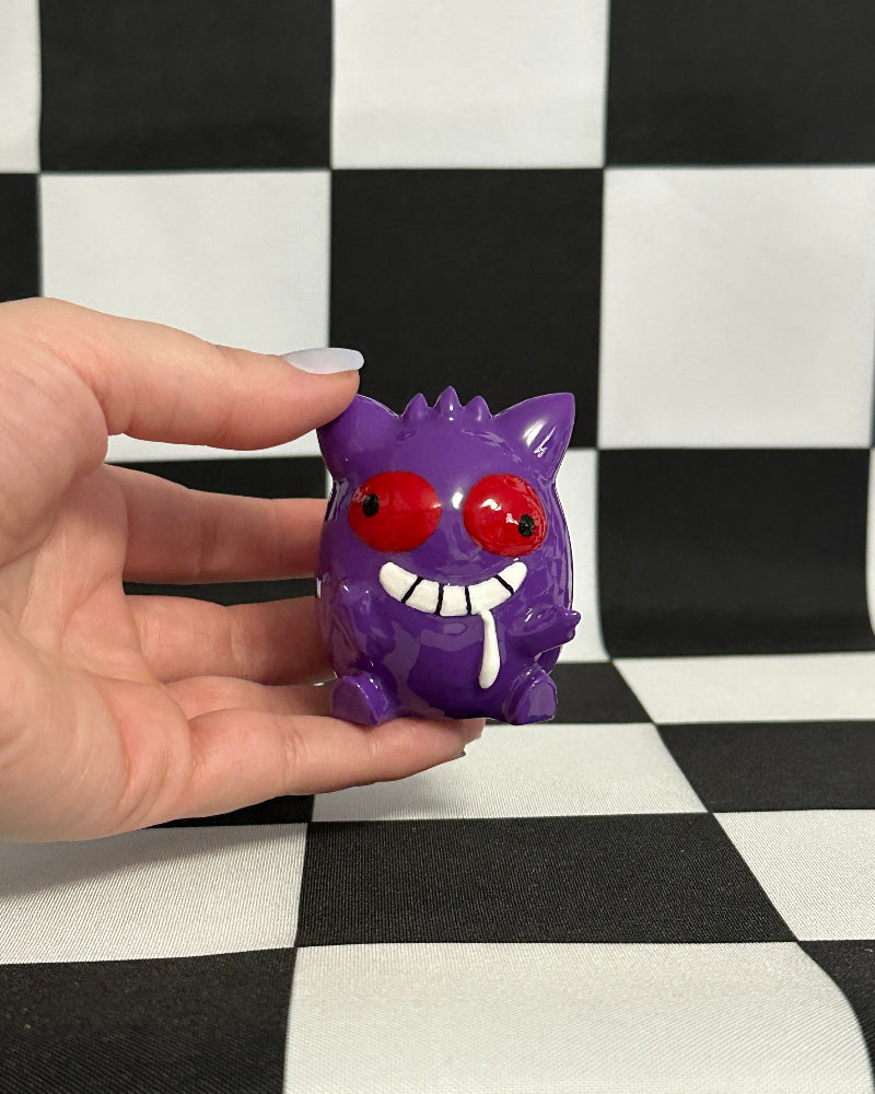 Hand holding a purple monster figurine with red eyes against a black and white checkered background