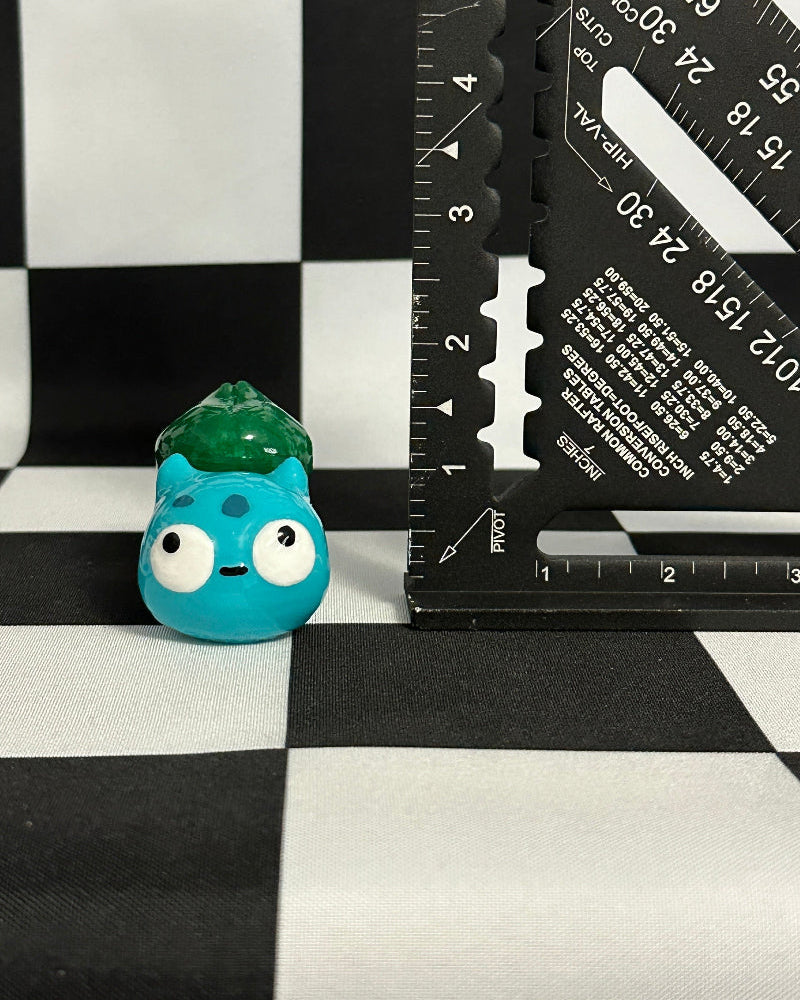 Small blue creature figurine with a green gem on a black and white checkered background with a ruler.