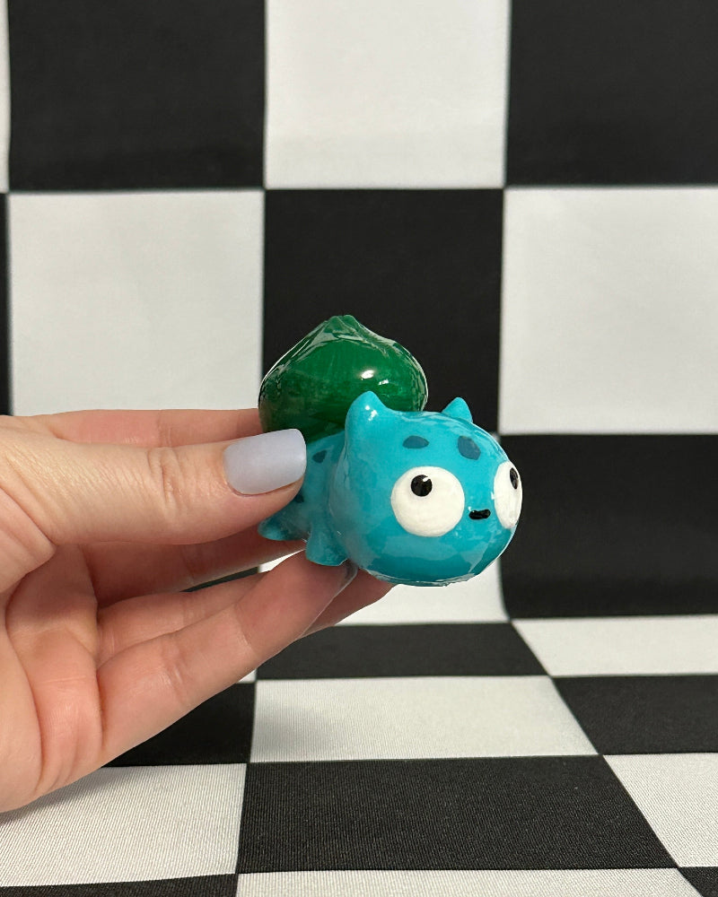 Hand holding a small blue and green creature toy on a black and white checkered background
