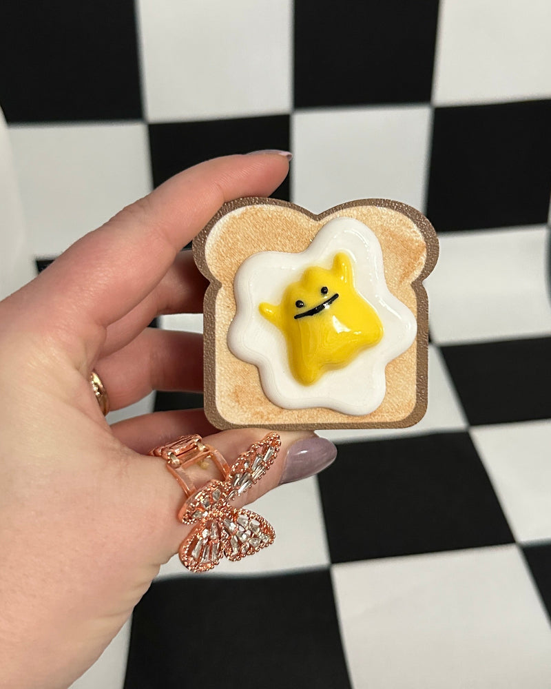 Hand holding a small plush toy shaped like a piece of toast with an egg on a checkered background