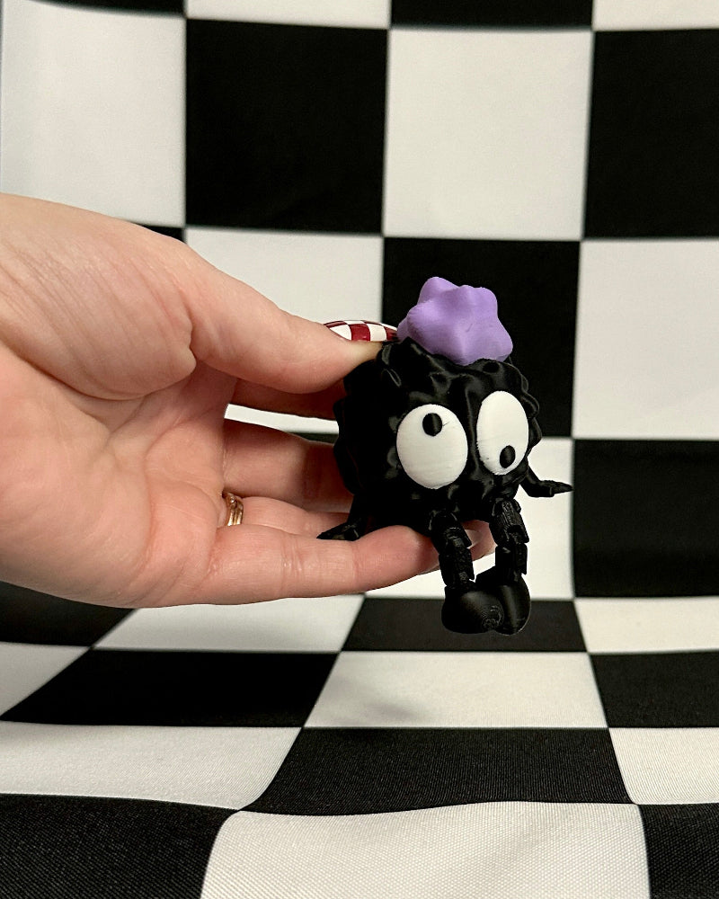 Hand holding a small black creature with googly eyes and purple hair against a checkered background