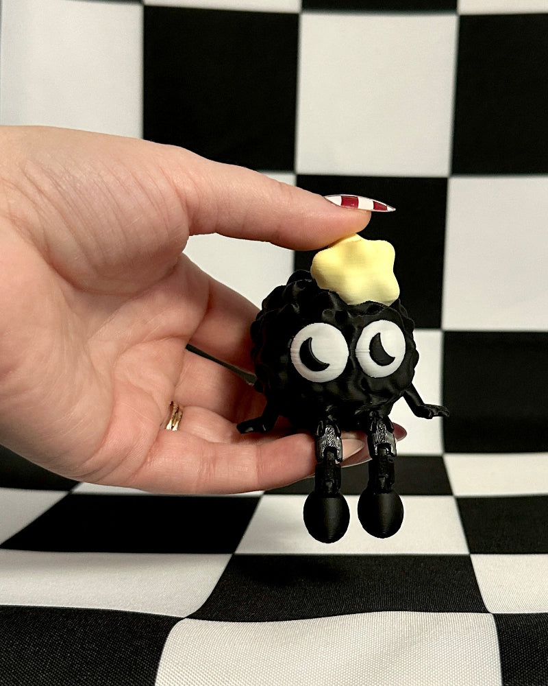 Hand holding a small black figurine with googly eyes on a black and white checkered background