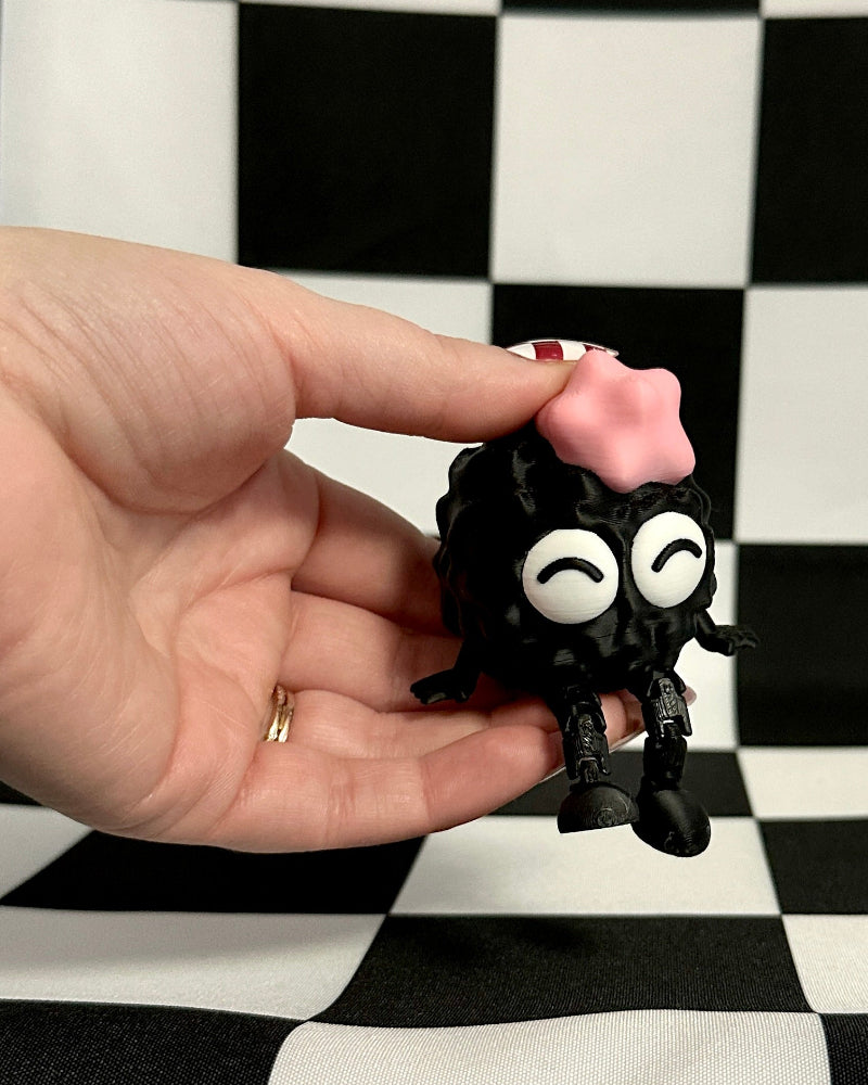 Hand holding a small black creature with googly eyes and purple hair against a checkered background