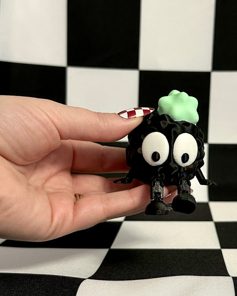 Small black creature with googly eyes held by a hand against a checkered background
