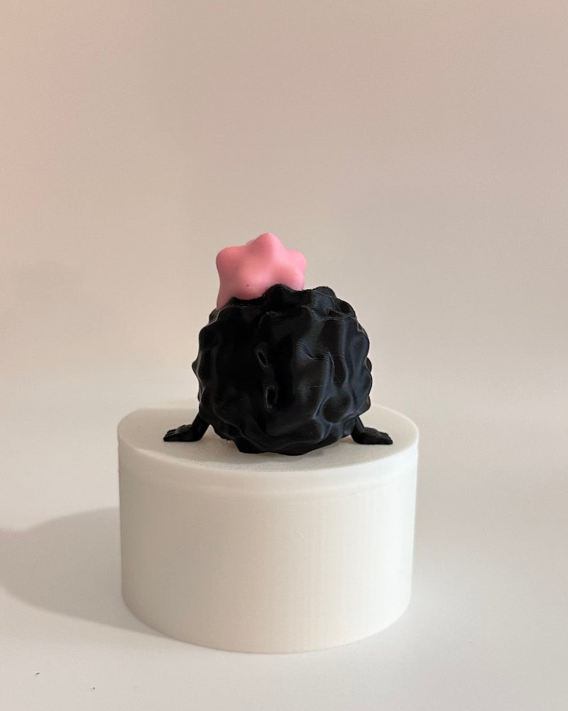Small black and pink figurine on a white cylindrical base against a neutral background