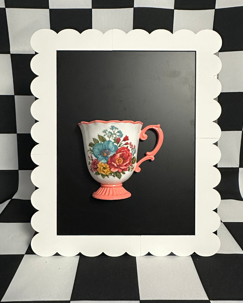 Decorative pink teacup with floral design on a black background framed by a white scalloped border against a black and white checkered wall.