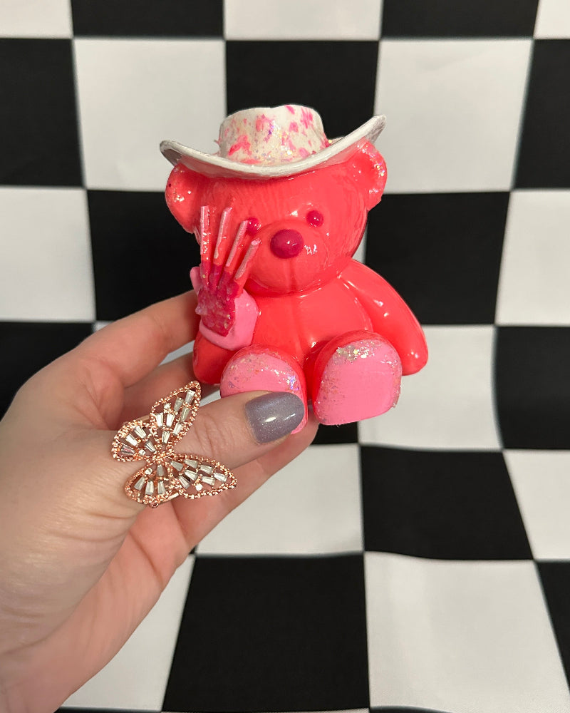 Pink teddy bear figurine with a cowboy hat held by a hand on a black and white checkered background