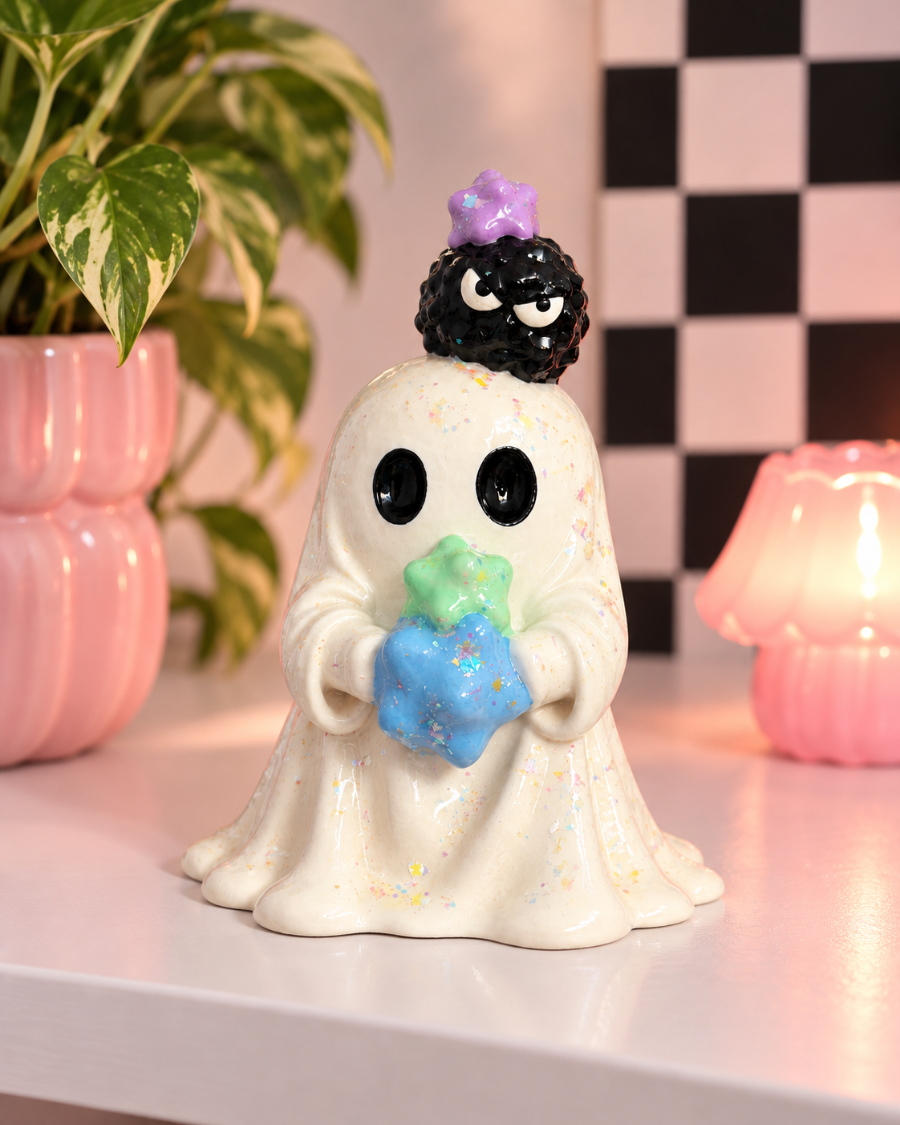 A ceramic ghost figurine with a white body, black eyes, a black spiky hat with a purple crown, and a blue and green star-shaped accessory, displayed on a tabletop with a plant and pink lamp in the background.