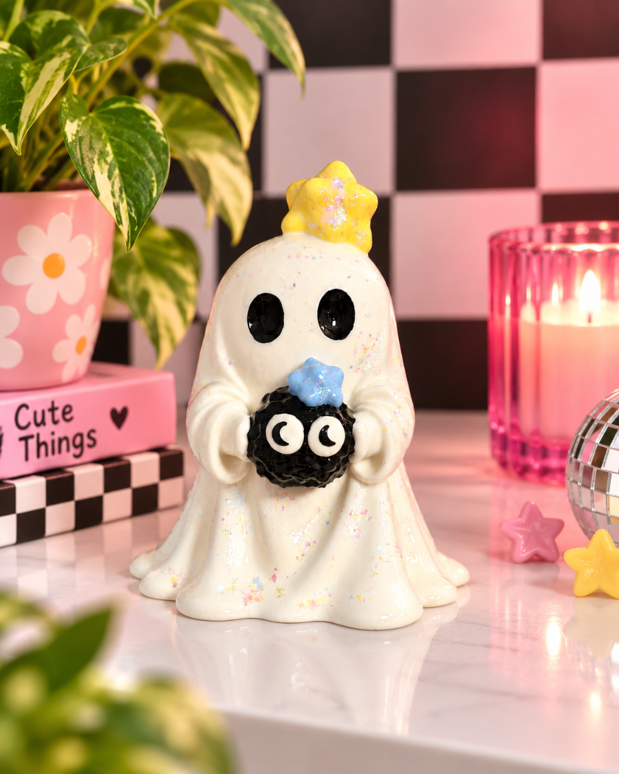 A cute ceramic ghost figurine holding a black spider, decorated with a yellow crown and blue bow, set on a tabletop with a pink flower pot, checkered background, and a lit candle.