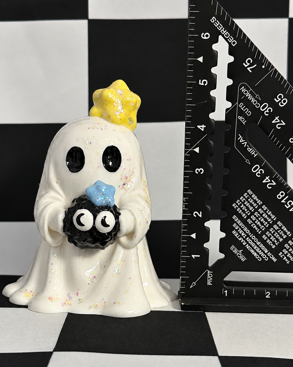 A small white ghost figurine with black eyes, holding a black spider with a blue bow, standing next to a black triangular ruler on a black-and-white checkered surface.