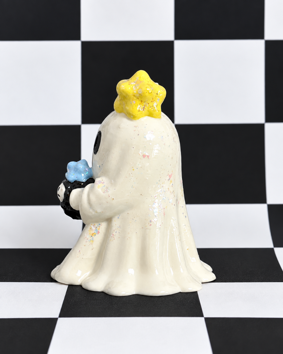 A ceramic ghost figurine with a yellow glittery star on its head, holding a small blue flower, set against a black and white checkered background.