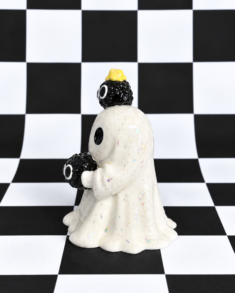A ghost-shaped figurine with a white glittery body, black eyes, and two black spherical creatures on its head and shoulders, one wearing a small yellow hat, set against a black and white checkered background.
