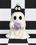 A ceramic ghost figurine holding a purple glittery flower, with a small black creature on its head wearing a green hat, set against a black and white checkered background.