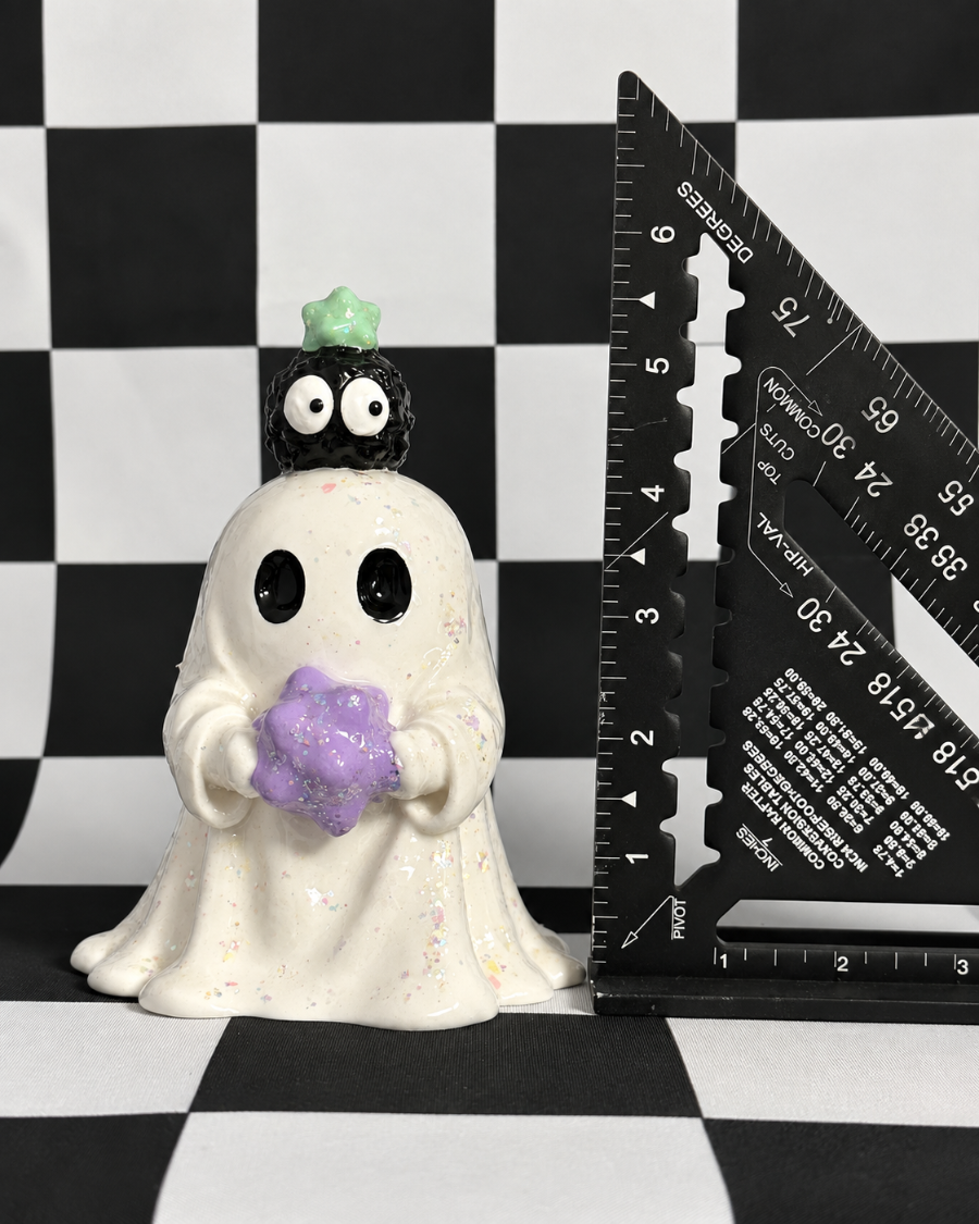 A ceramic Halloween ghost figurine with a white body, black eyes, a small black skull on its head, and a purple glittery pumpkin, shown next to a black triangular ruler on a black-and-white checkered background.