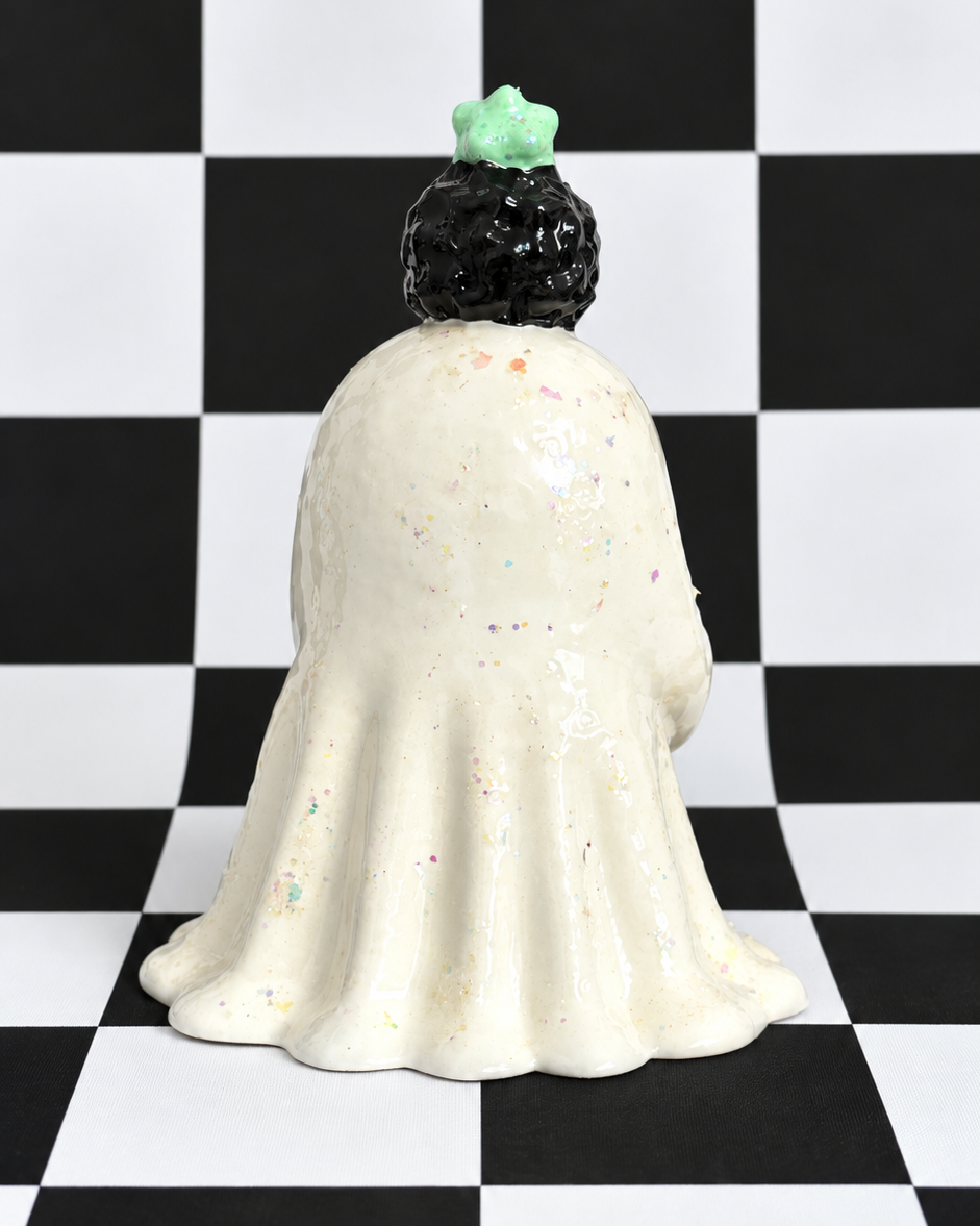 A ceramic ghost figurine shown from the back, featuring a cream-colored body with subtle multicolor speckles, a black textured hair bun, and a small mint green crown, set against a black and white checkered background.