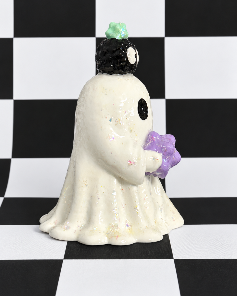 A small ceramic ghost figurine with a white body, black eyes, a black hat with a green pom-pom, and a purple star, standing on a black and white checkered background.