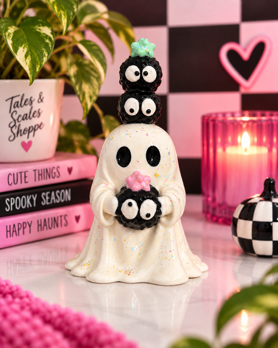 A Halloween-themed ghost figurine decorated with three stacked black cupcakes topped with green and pink frosting, holding a matching black cupcake in its hands. The figurine is styled on a reflective white surface with pink books, a plant, a pink candle, and a checkered background.